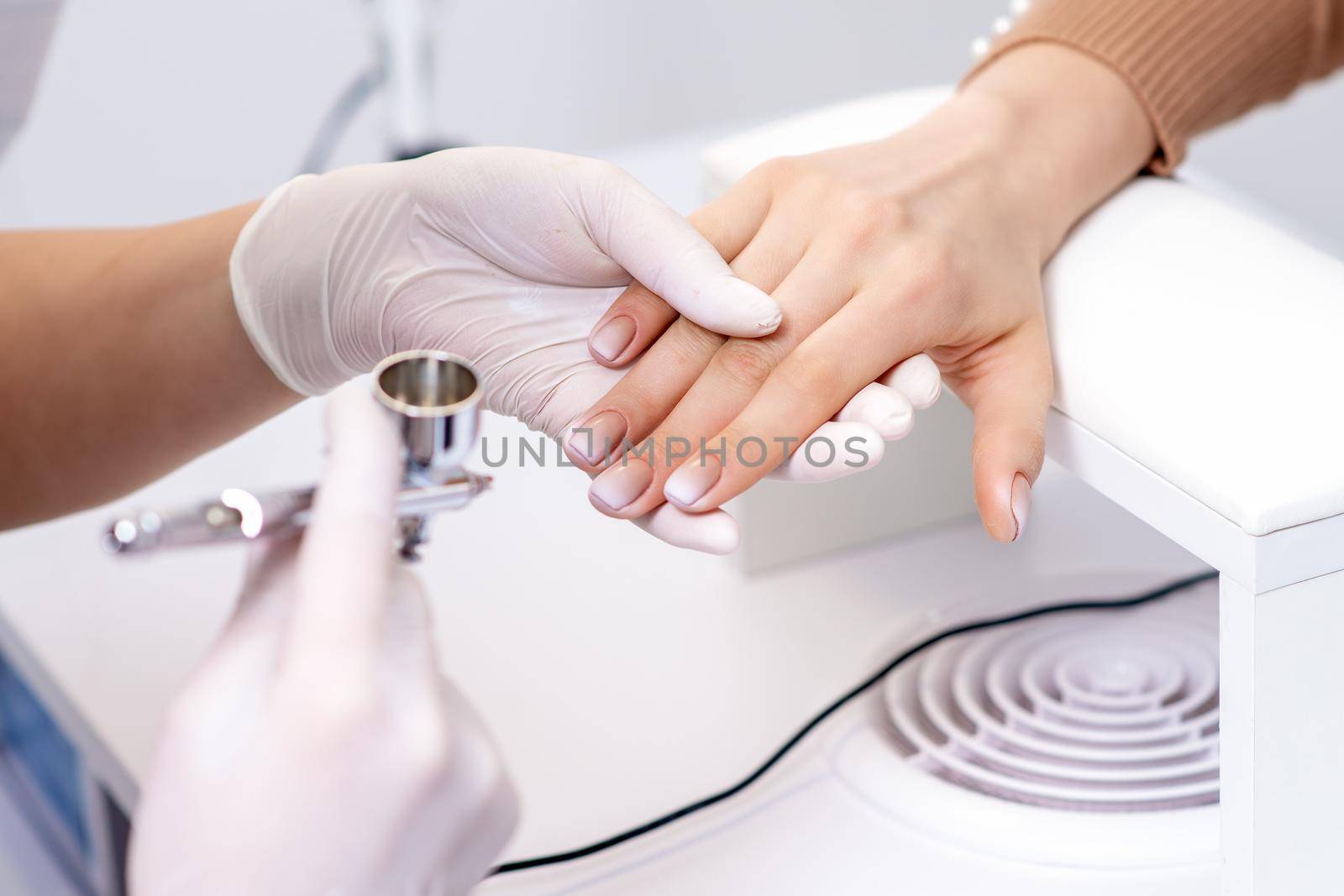 Young woman receiving manicure by airbrush in nail salon. Procedure for spraying paint on the nails
