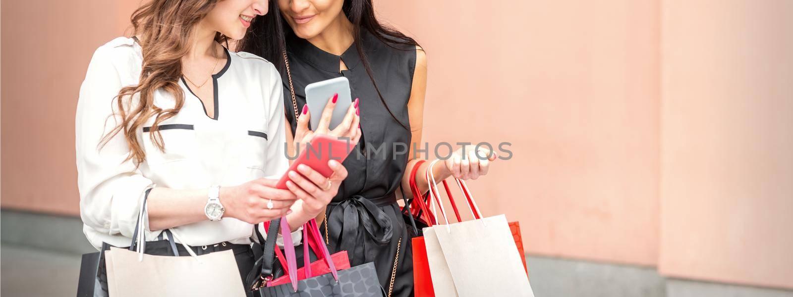Two beautiful caucasian young women watching at smart phone on new seasonal offers with bags during shopping near wall of shopping mall