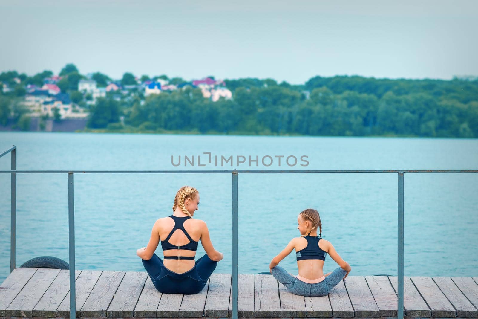 Mother and daughter doing gym exercises on the grass at the pier of the river