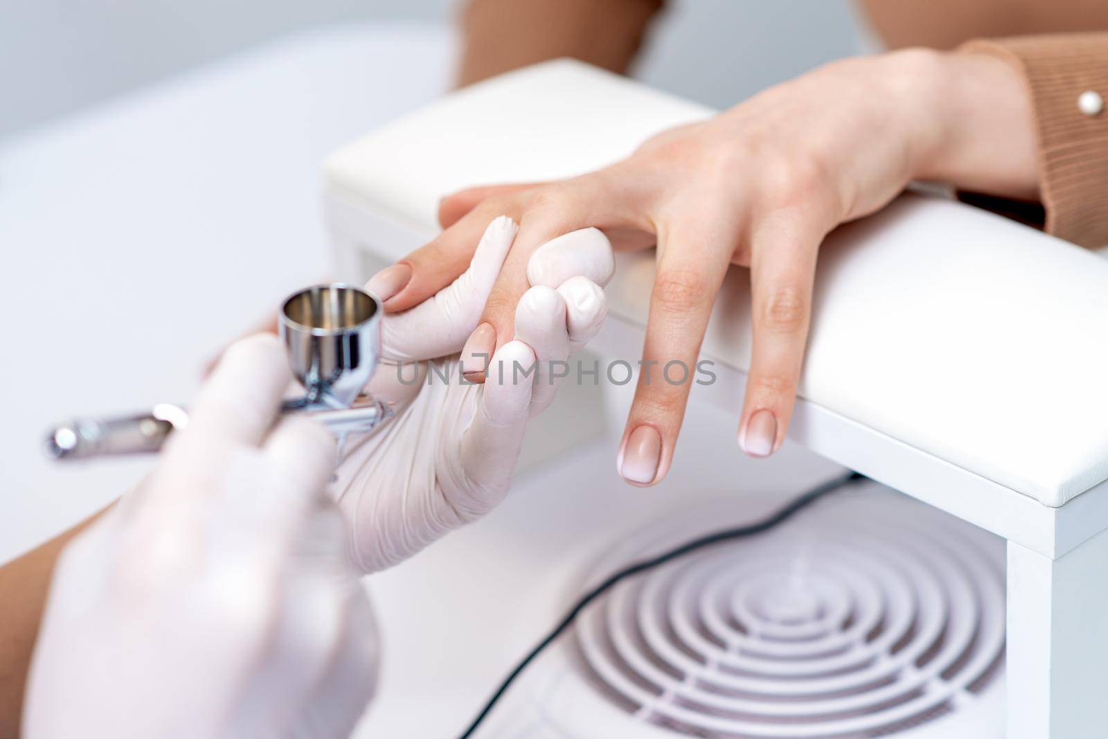 Young woman receiving manicure by airbrush in nail salon. Procedure for spraying paint on the nails
