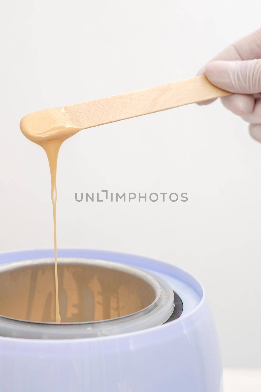 Wax heater with hot yellow wax on wooden spatula in human hand. Hot wax for depilation. Salon equipment