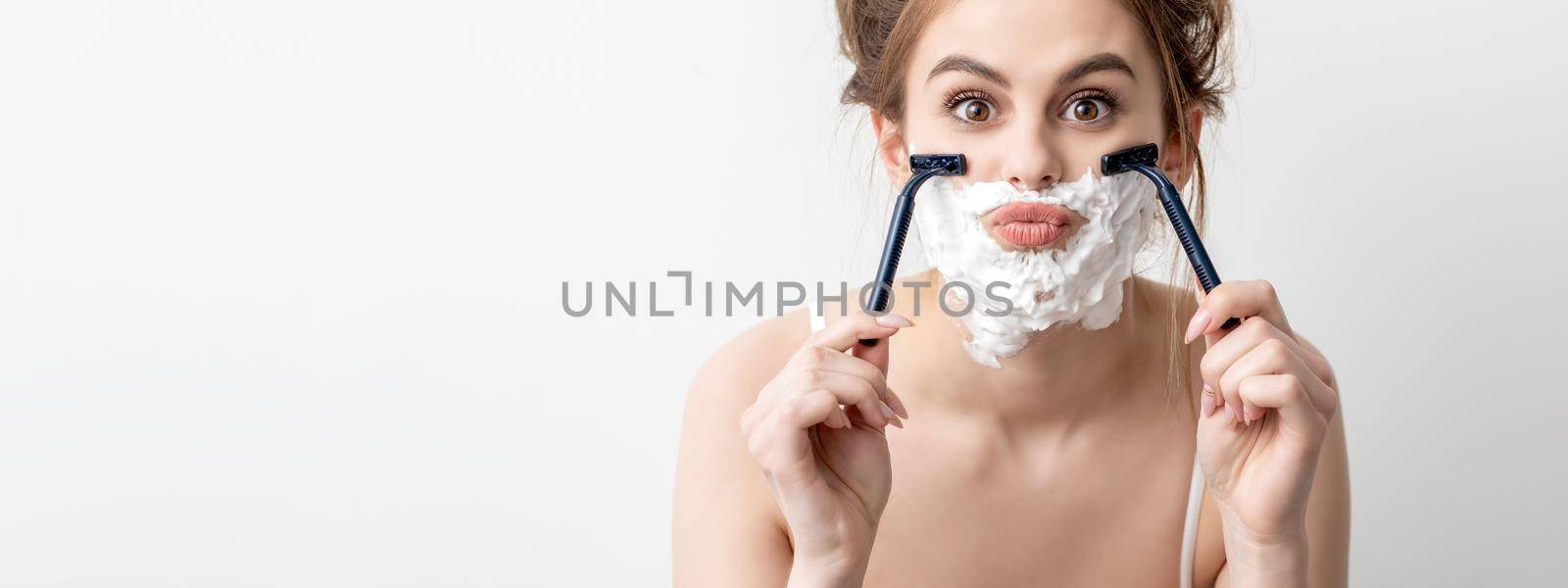 Beautiful young caucasian woman shaving her face by two razors on white background. Pretty woman with shaving foam on her face