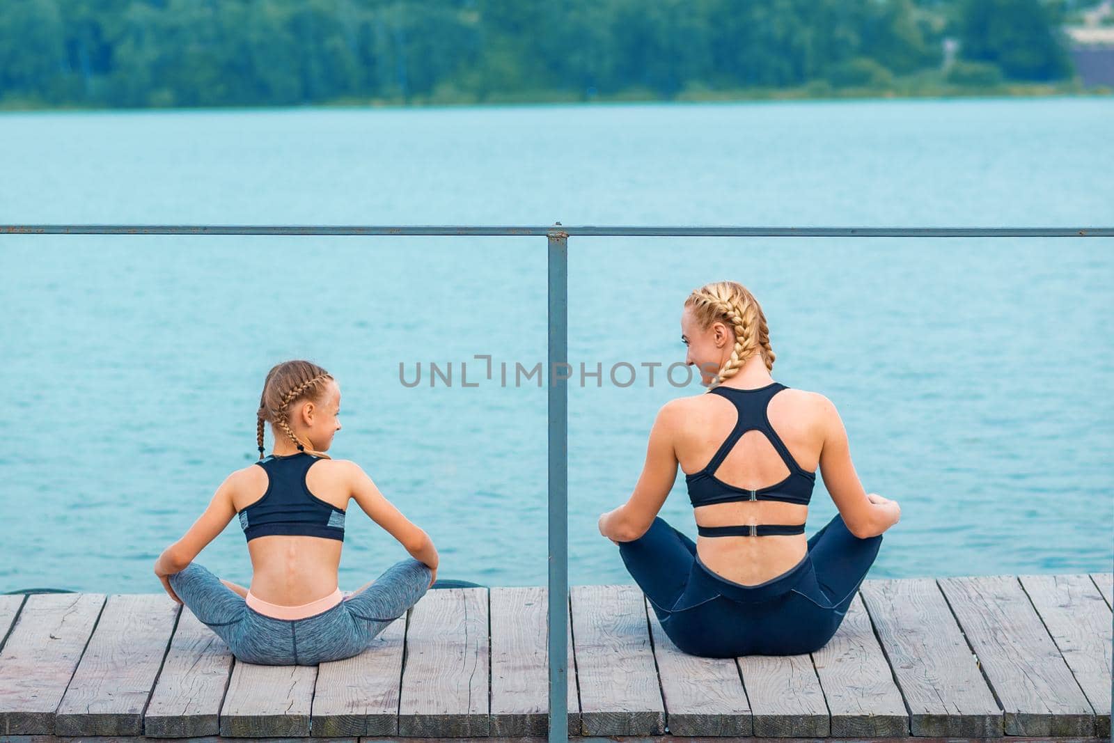 Mother and daughter doing gym exercises on the grass at the pier of the river