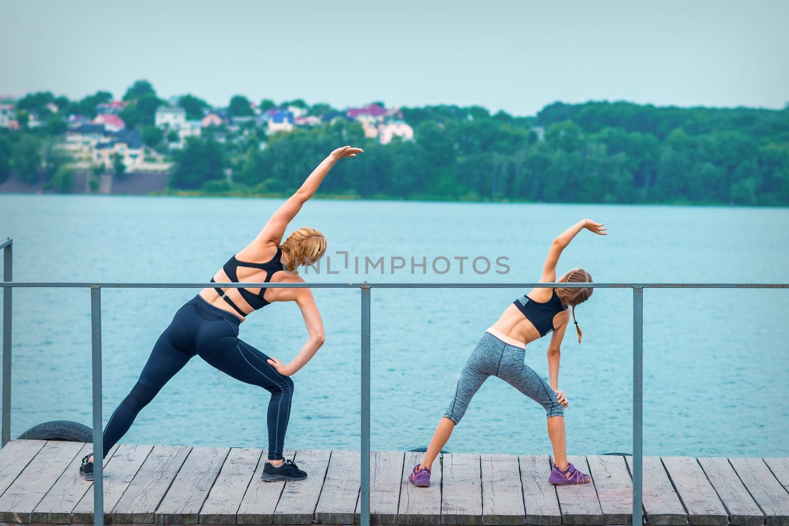 Mother and daughter doing gym exercises on the grass at the pier of the river