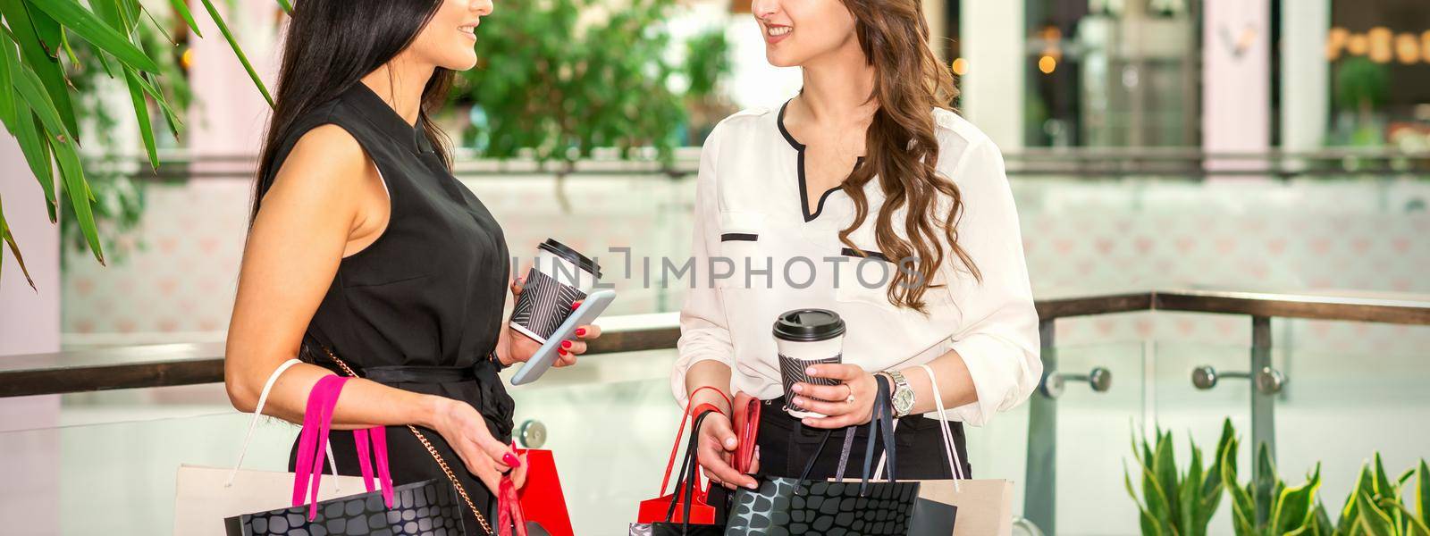 Two beautiful happy young women with shopping bags and cups of coffee discussing purchases looking at each other and smiling at shopping mall