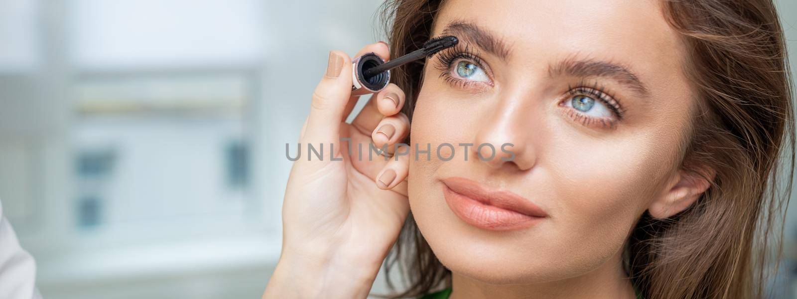 Professional makeup artist applying mascara on lashes of beautiful young caucasian woman in beauty salon