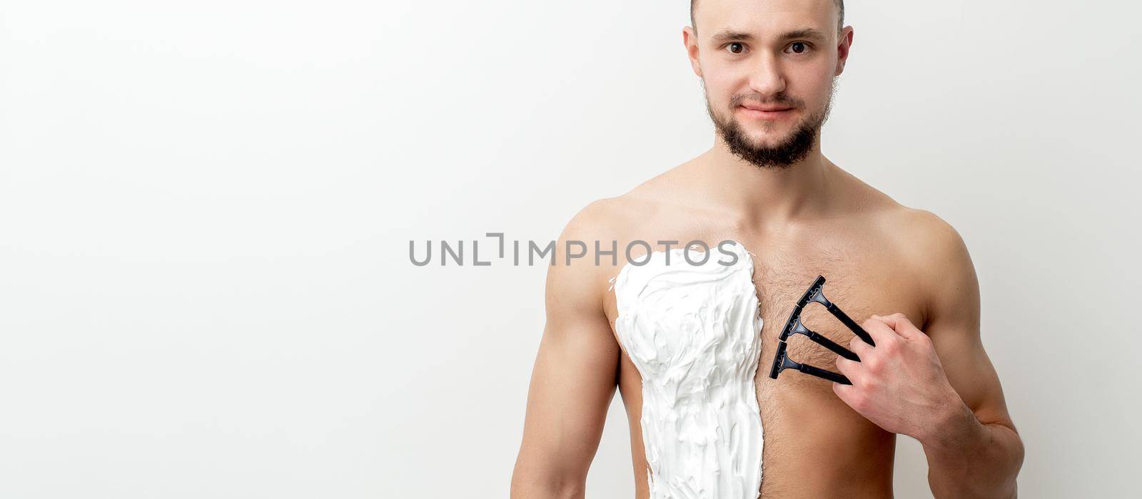 Young caucasian man with beard holds razor shaves his chest with white shaving foam on white background. Man shaving his torso