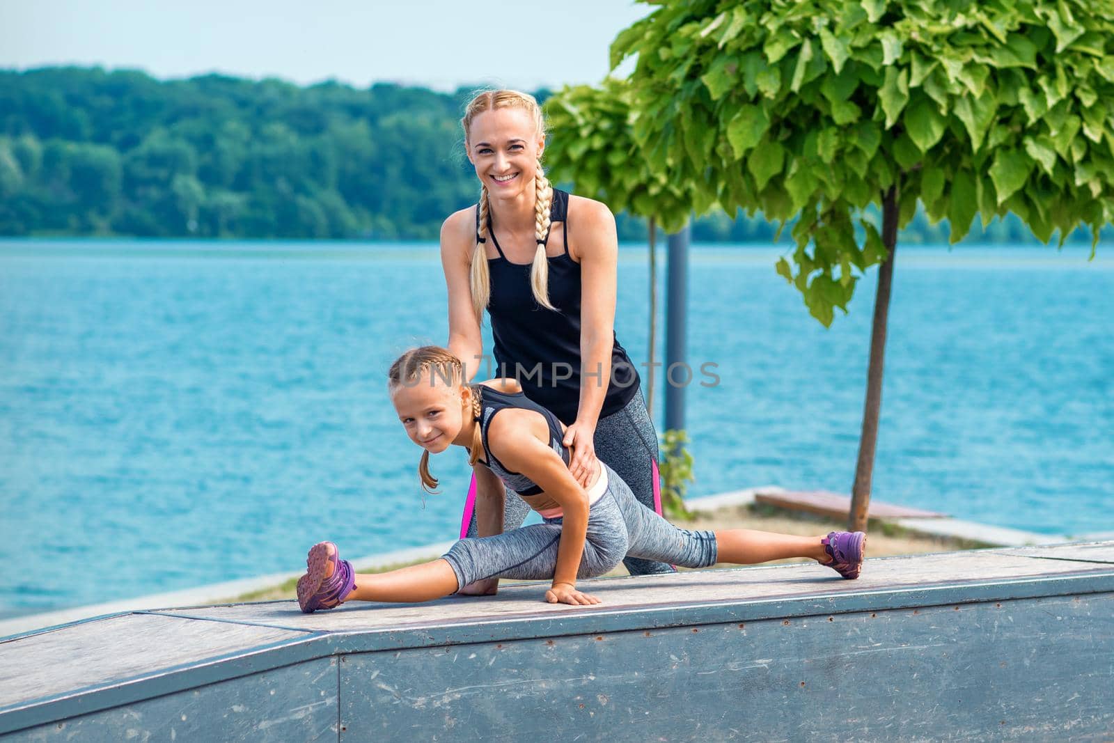 Mother and daughter doing gym exercises on the grass at the pier of the river