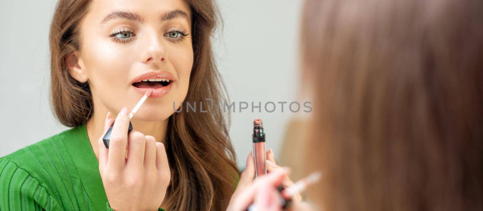 Beautiful young caucasian woman applying gloss to the lips looking in the mirror