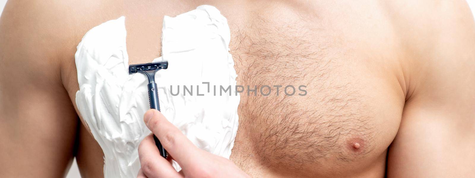 Young caucasian man with beard holds razor shaves his chest with white shaving foam on white background. Man shaving his torso