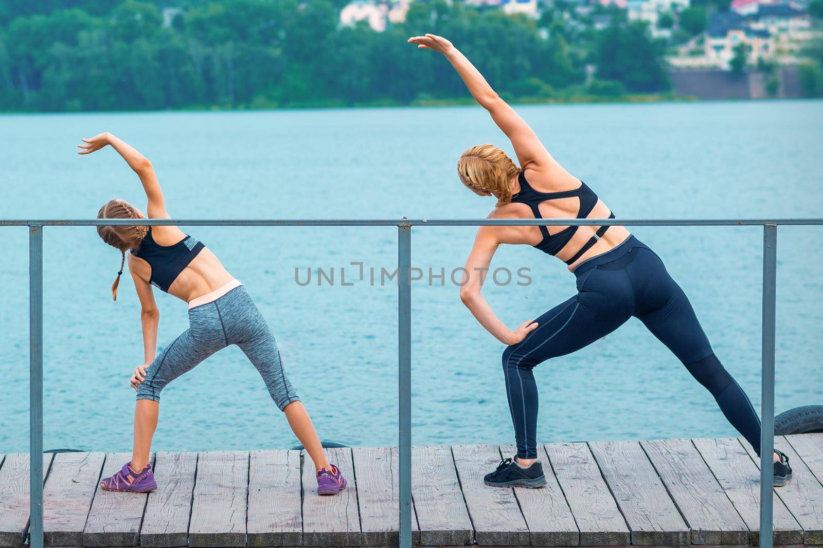 Mother and daughter doing gym exercises on the grass at the pier of the river