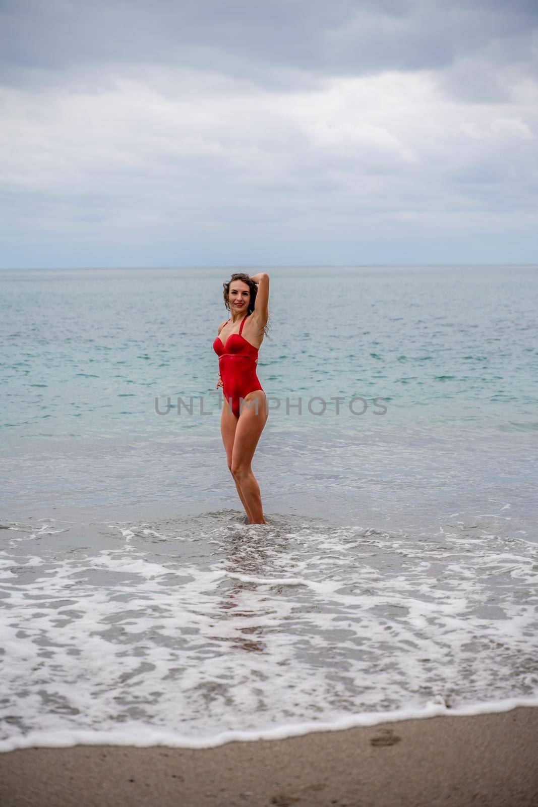 A beautiful and sexy brunette in a red swimsuit on a pebble beach, Running along the shore in the foam of the waves by Matiunina