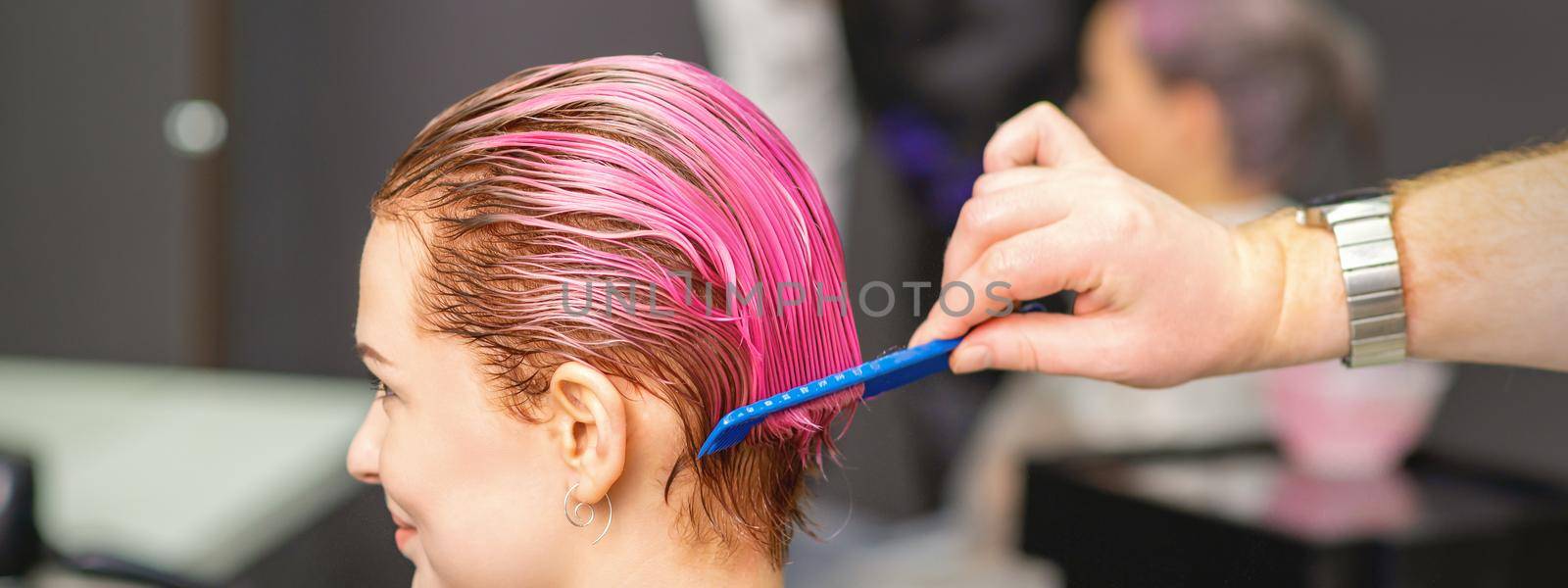 Young woman receiving hair treatment after pink coloring by hand of male hairdresser in hair salon