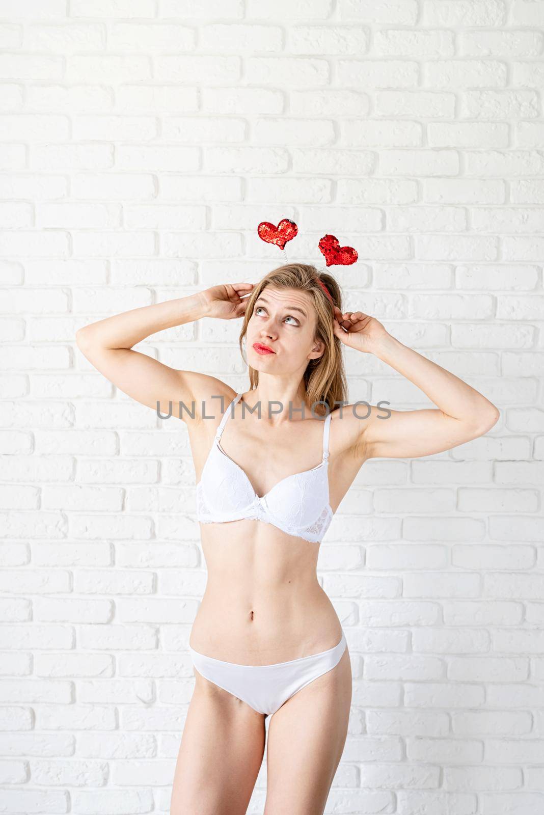 Full length of attractive young brown hair woman in white underwear wearing heart shaped headband posing against white brick wall background