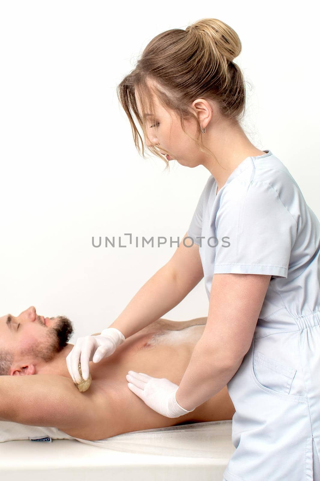 Young man receiving waxing underarm or epilation armpit by young female cosmetologist in beauty salon
