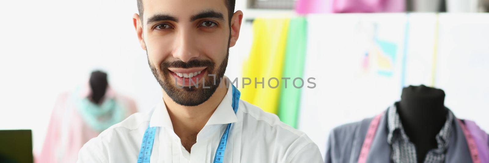 Portrait of charming young man wear tape measure on neck and hold scissors. Mannequin in atelier on background, talented tailor. Develop business concept
