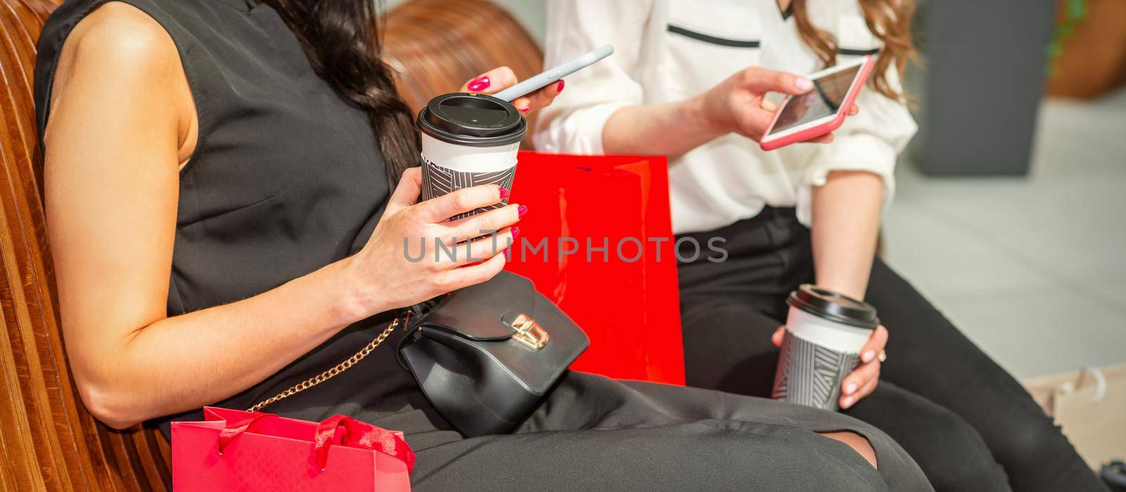 Two beautiful caucasian young women looking in smart phone with bags sitting on the bench while resting in the shopping mall