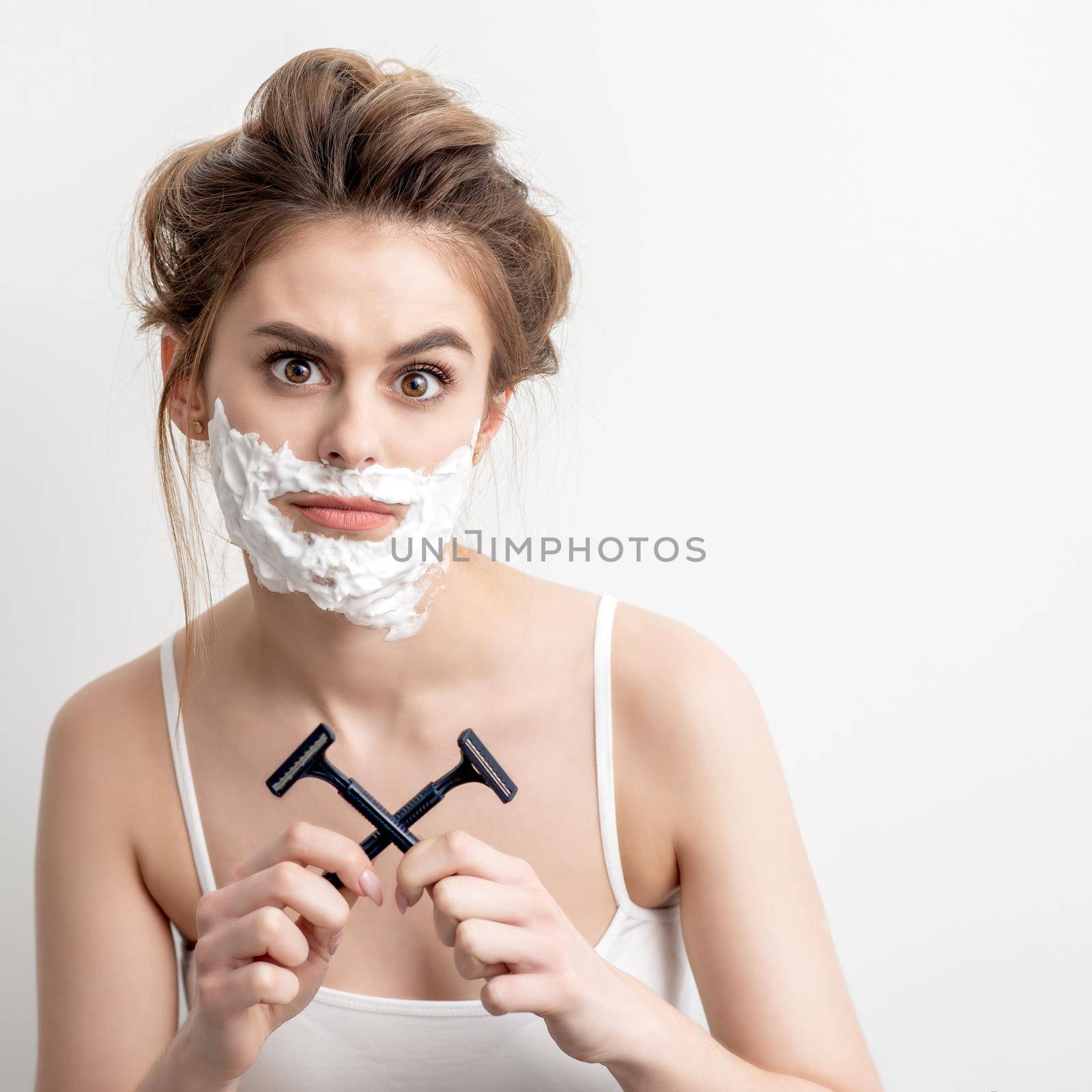Beautiful young caucasian woman with shaving foam on her face and two razors in her hands on white background
