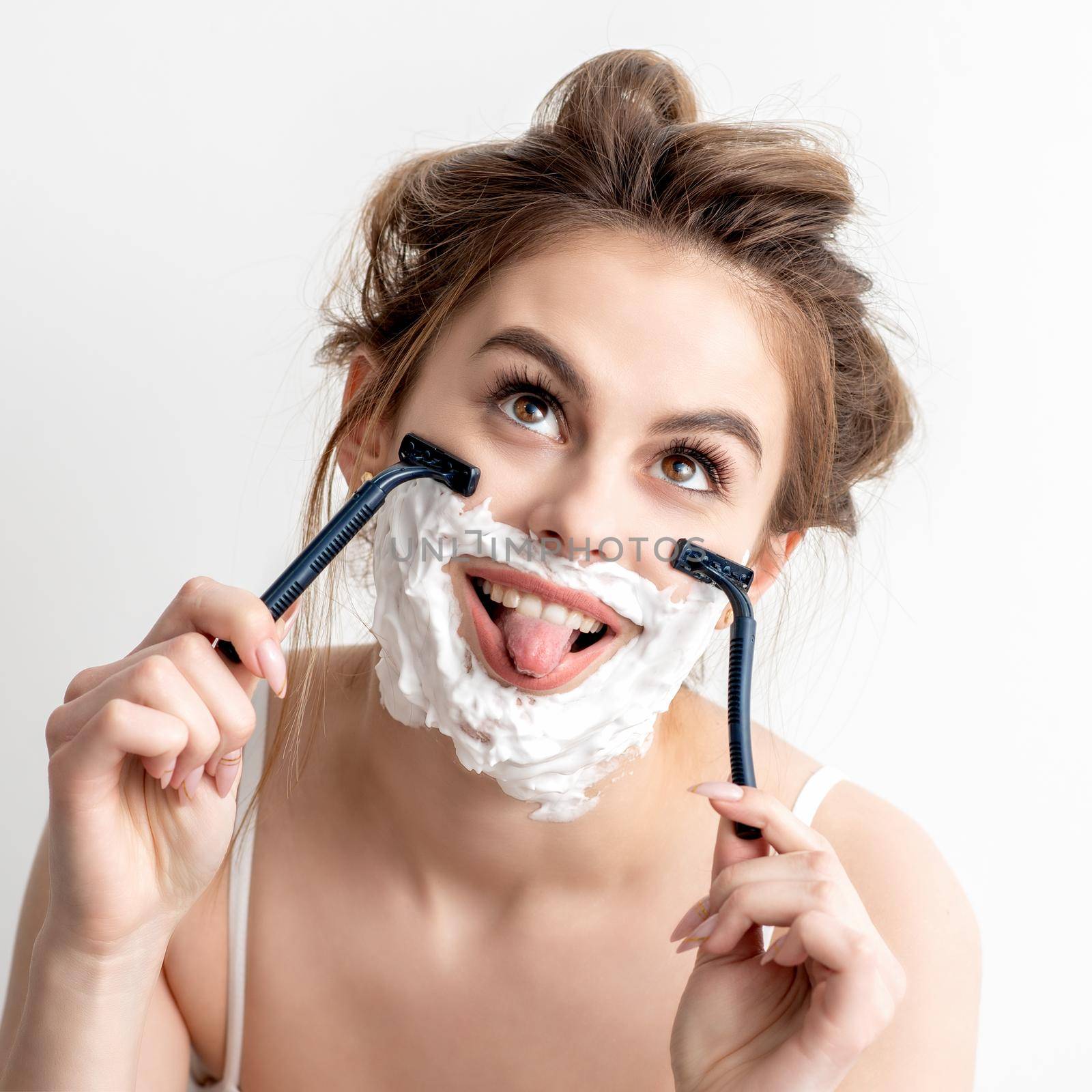 Beautiful young caucasian woman shaving her face by two razors with tongue out on white background.