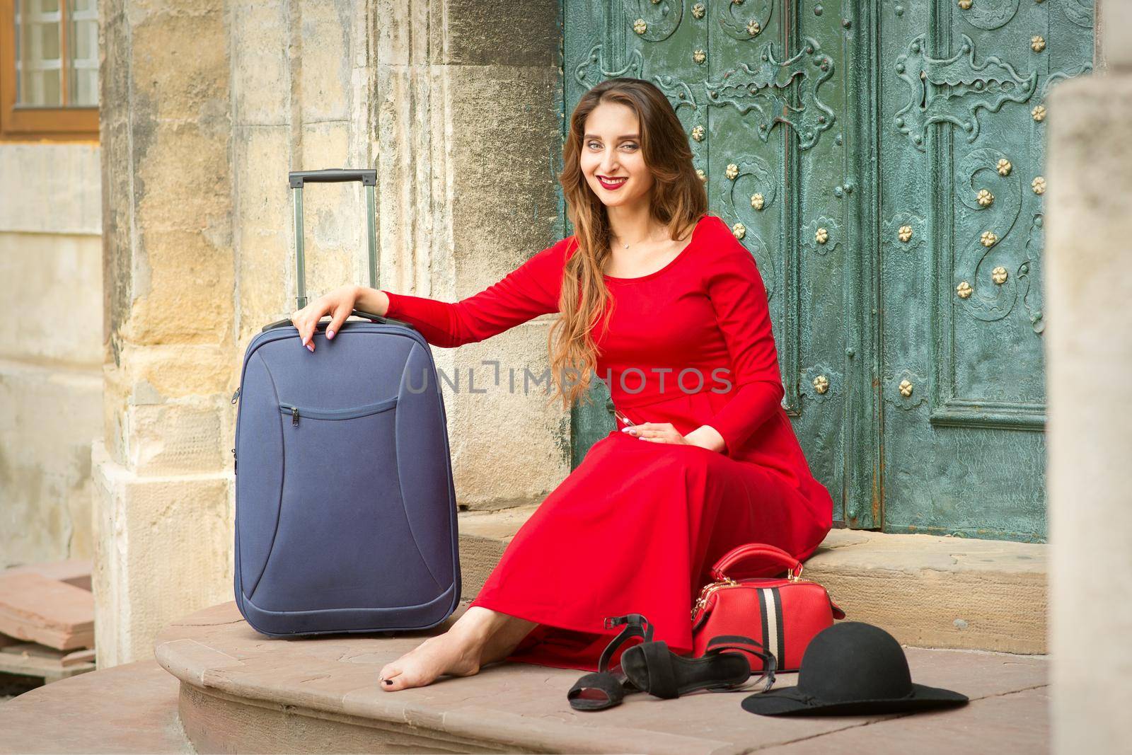Beautiful young caucasian woman sitting on the stairs by the door wearing black hat and long red dress with travel suitcase and smartphone