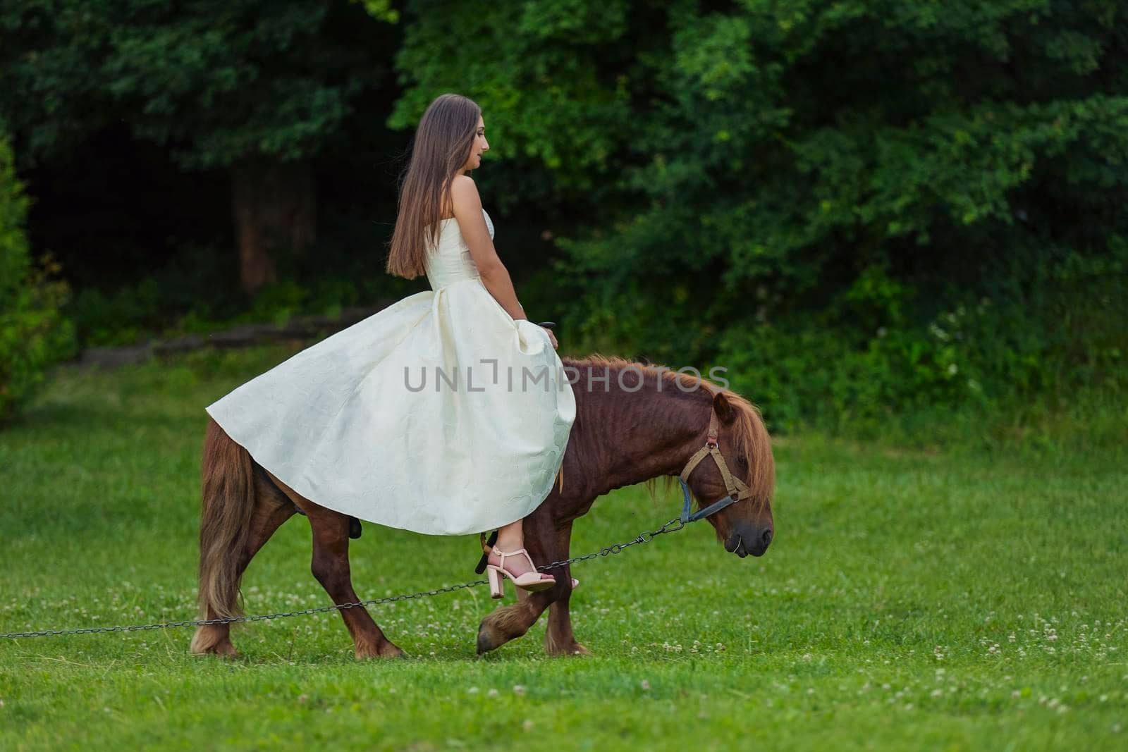 girl in a white dress rides a pony on a green lawn