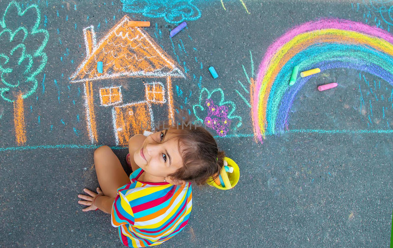 The child draws a house and a rainbow on the asphalt with chalk. Selective focus. by yanadjana