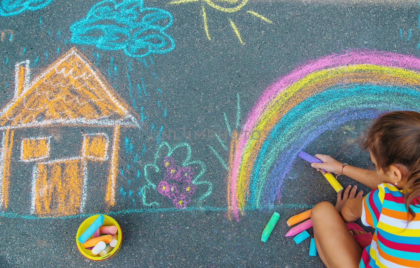 The child draws a house and a rainbow on the asphalt with chalk. Selective focus. Kids.