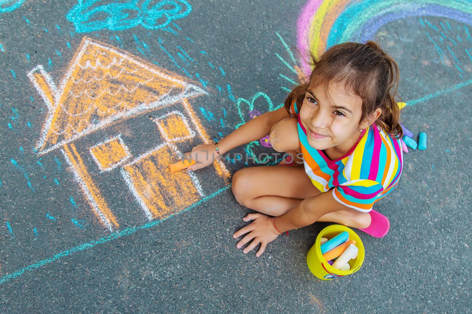 The child draws a house and a rainbow on the asphalt with chalk. Selective focus. Kids.