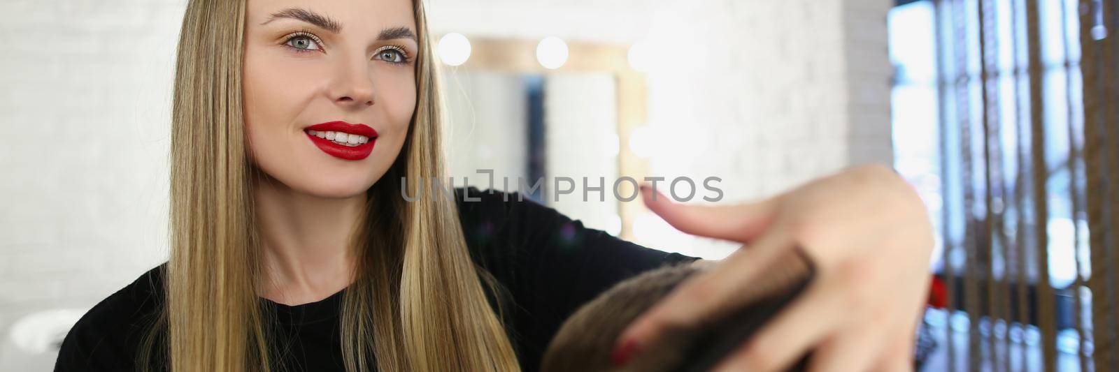 Portrait of beautiful young hair master brushing clients hair with tool and cut. Talented hairdresser work in beauty studio. Fashion, barbershop concept