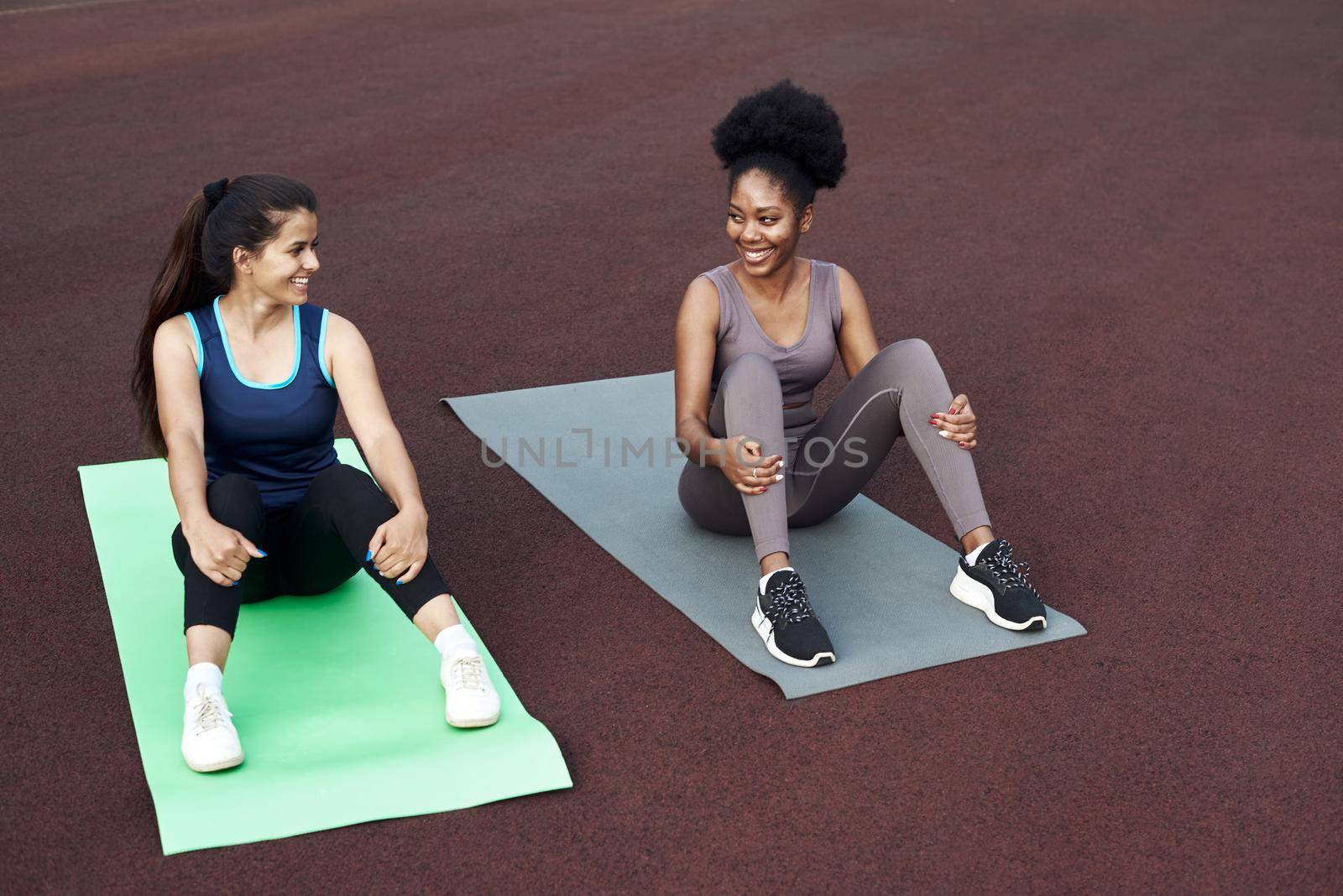 Break in training. Two diverse young women are sitting on yoga mats in the stadium and talking, laughing