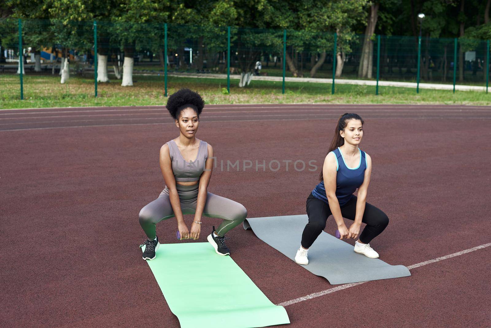 Young diverse friends squat at the stadium, warm up before performing group exercises.