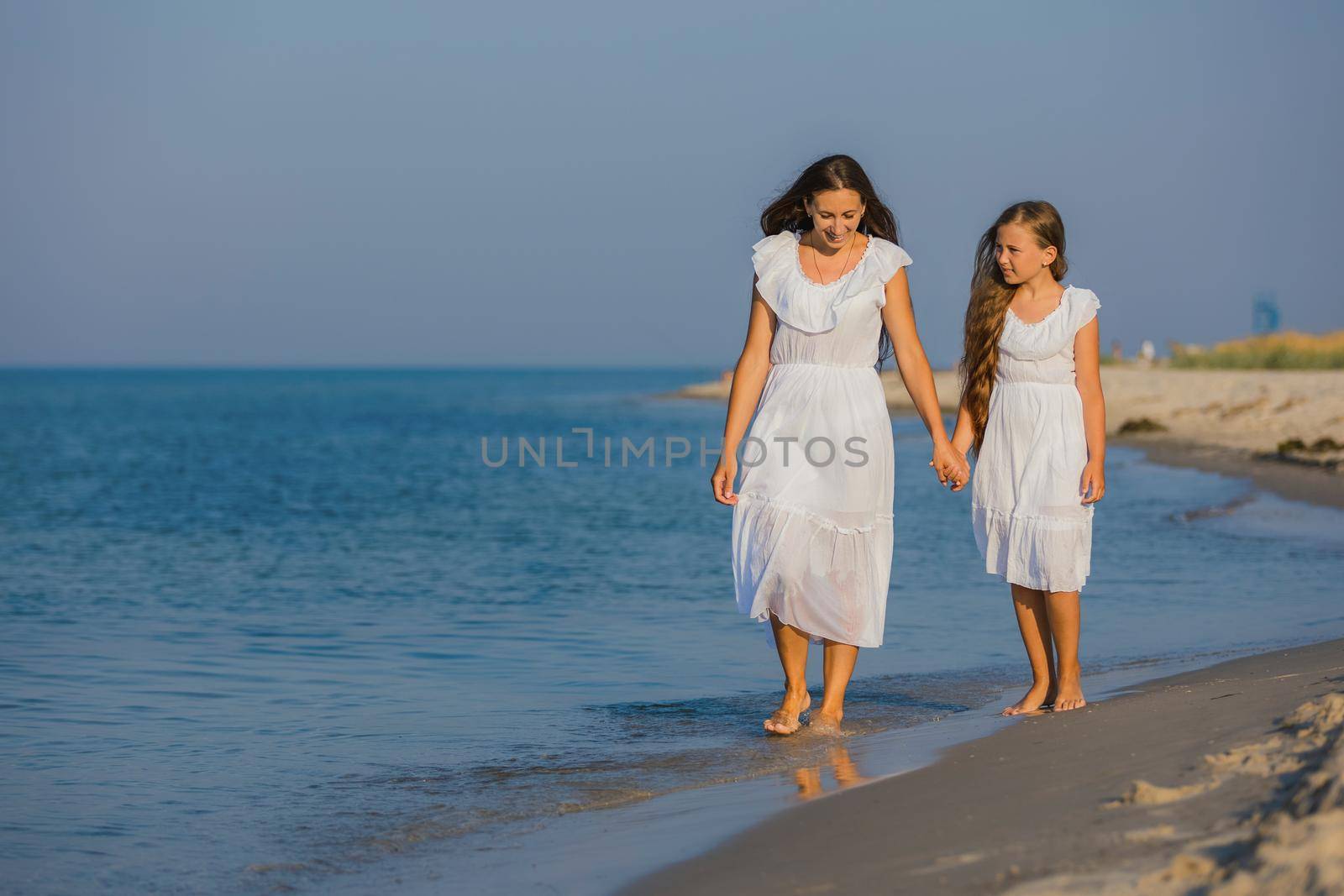 mom and daughter in white dresses walk on the beach