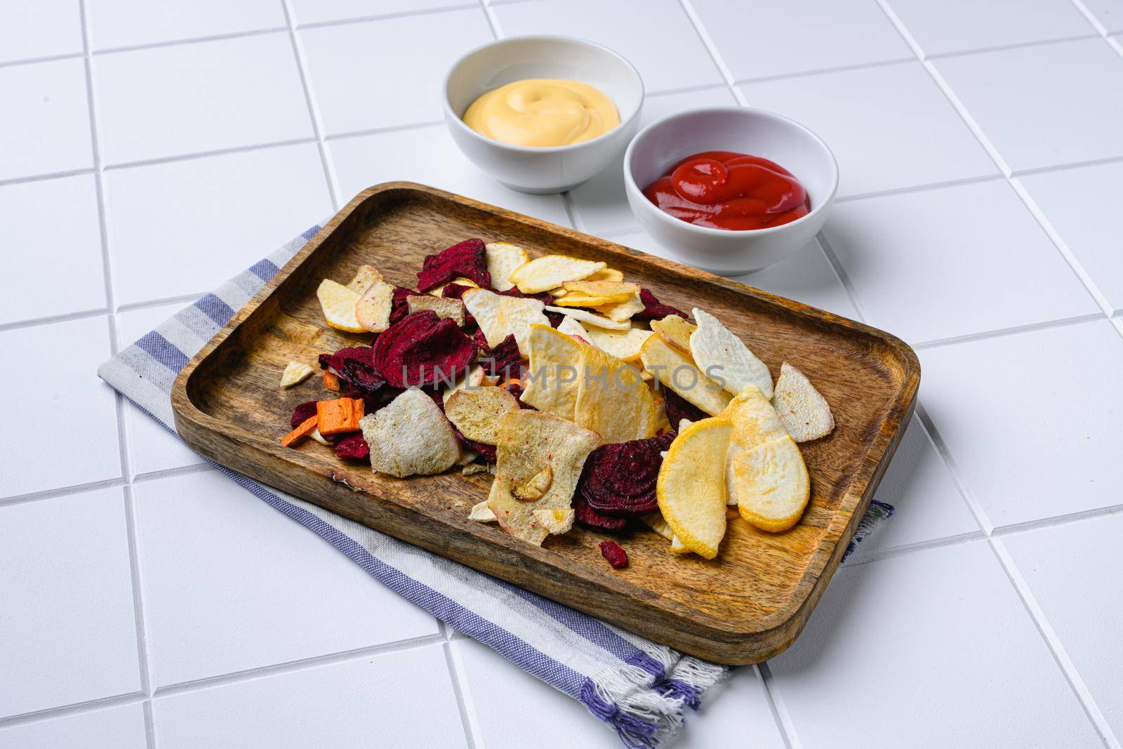 Dried vegetables chips from carrot, beet, parsnip and other vegetables, on white ceramic squared tile table background, with copy space for text by Ilianesolenyi