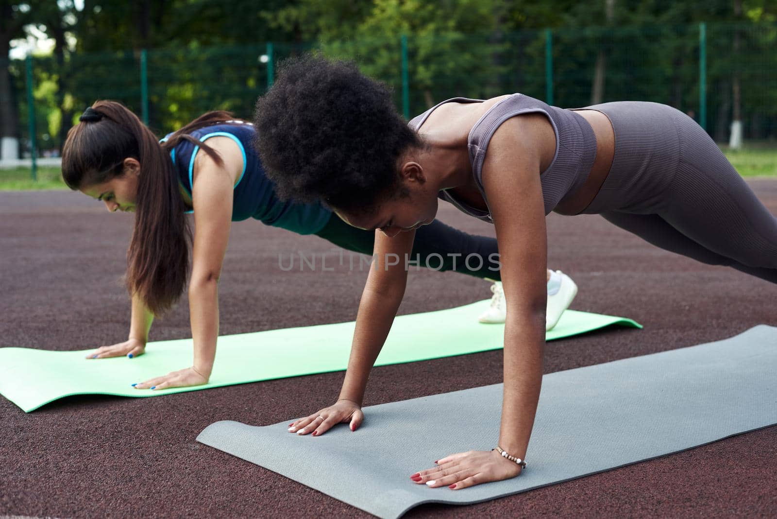 Two fitness women of different nationalities in a plank position in an outdoor stadium. Push ups