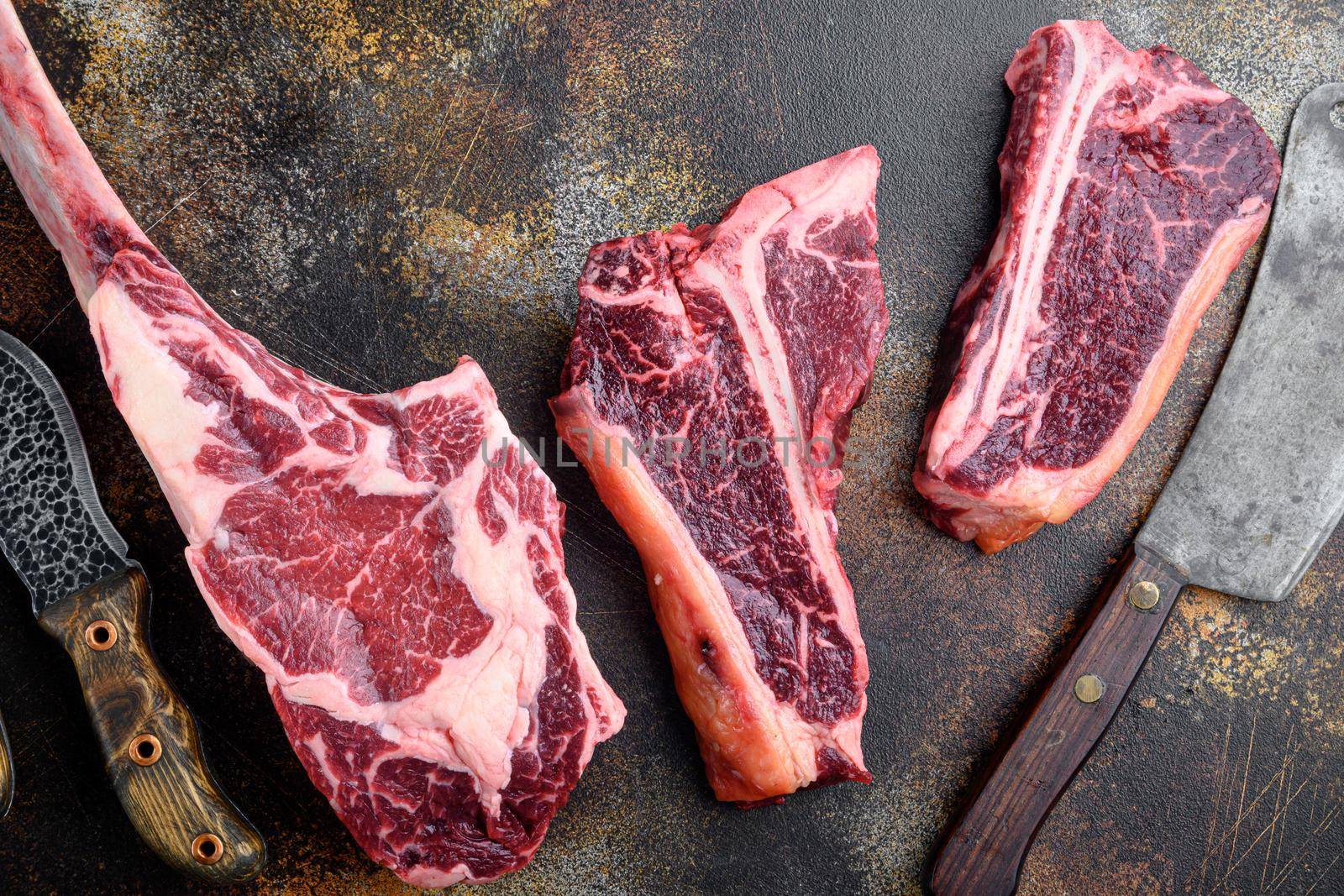 Raw prime beef meat dry aged steak set, tomahawk, t bone or porterhouse and club steak , with old butcher cleaver knife, on old dark rustic background, top view flat lay