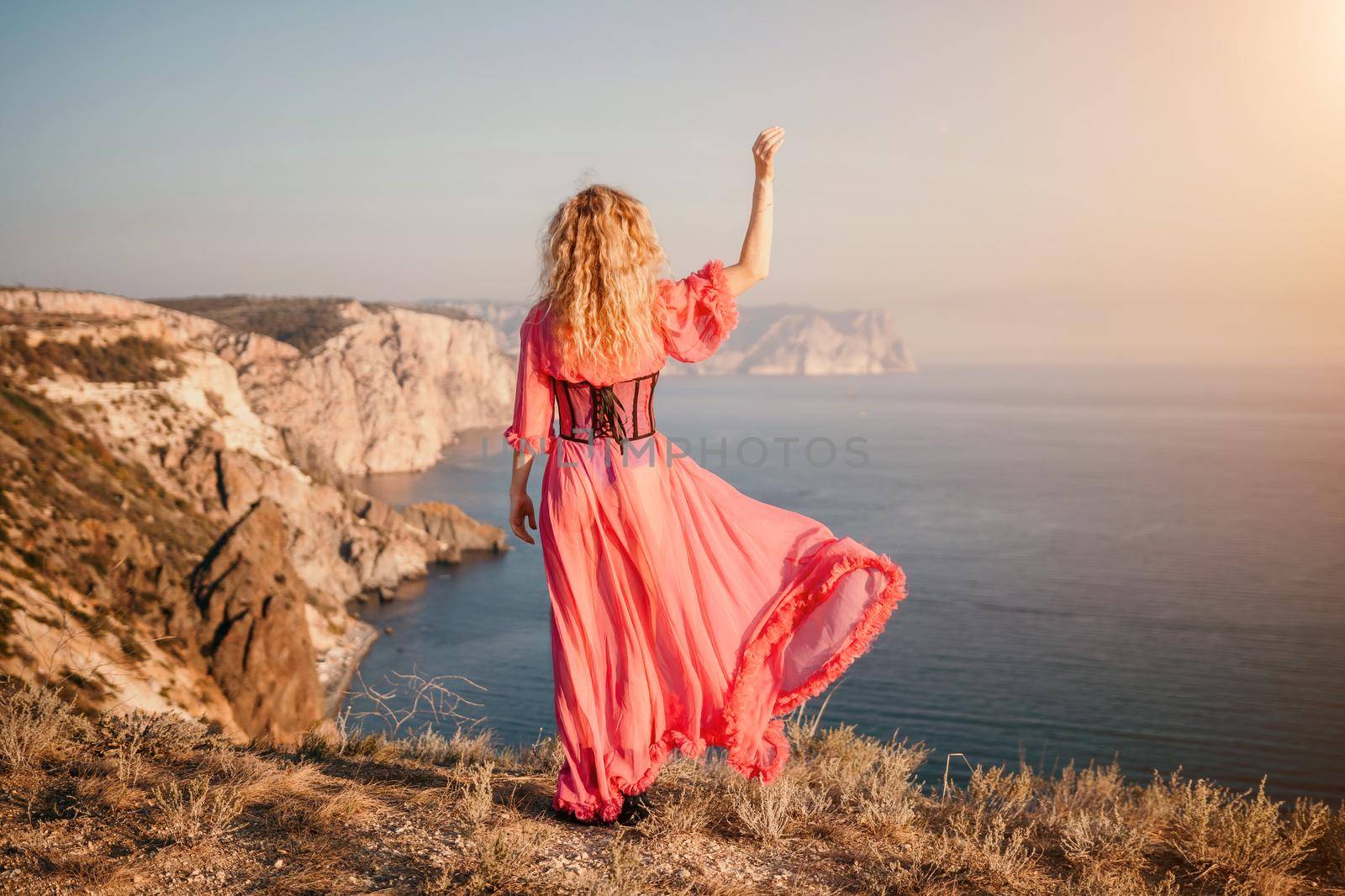 Beautiful young caucasian woman with curly blond hair and freckles. Cute redhead woman portrait in a pink long dress posing on a volcanic rock high above the sea during sunset. by panophotograph