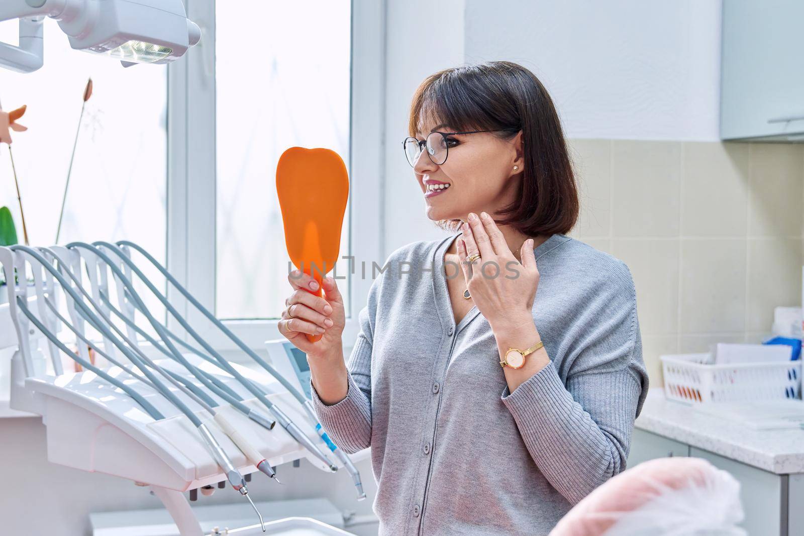 Happy middle aged woman dentist patient with a mirror in her hands looking at her teeth. Treatment, whitening, implants, medicine, health care and beauty smile concept