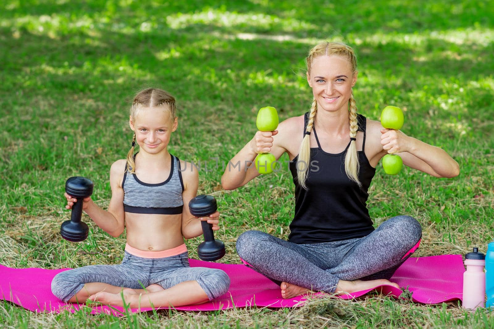 Young woman and girl are training with dumbbells sitting on the grass in the park