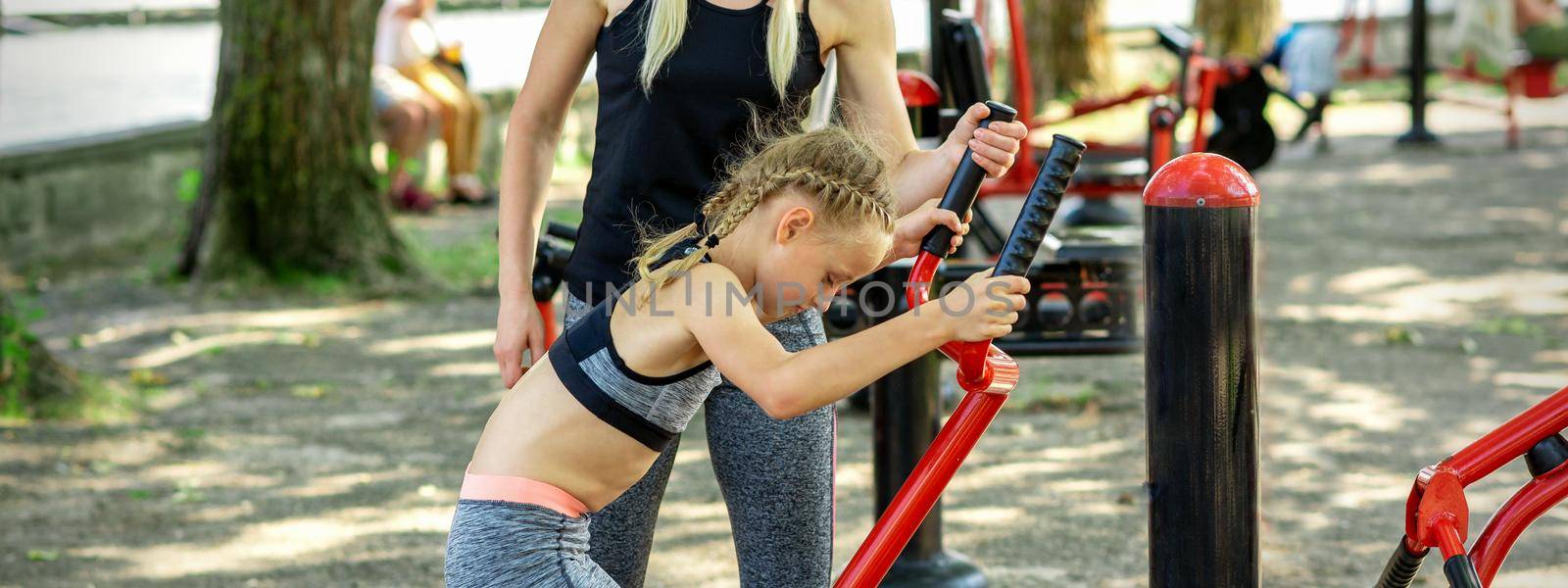 Little girl does exercises on simulator under supervision of young woman coach in the park