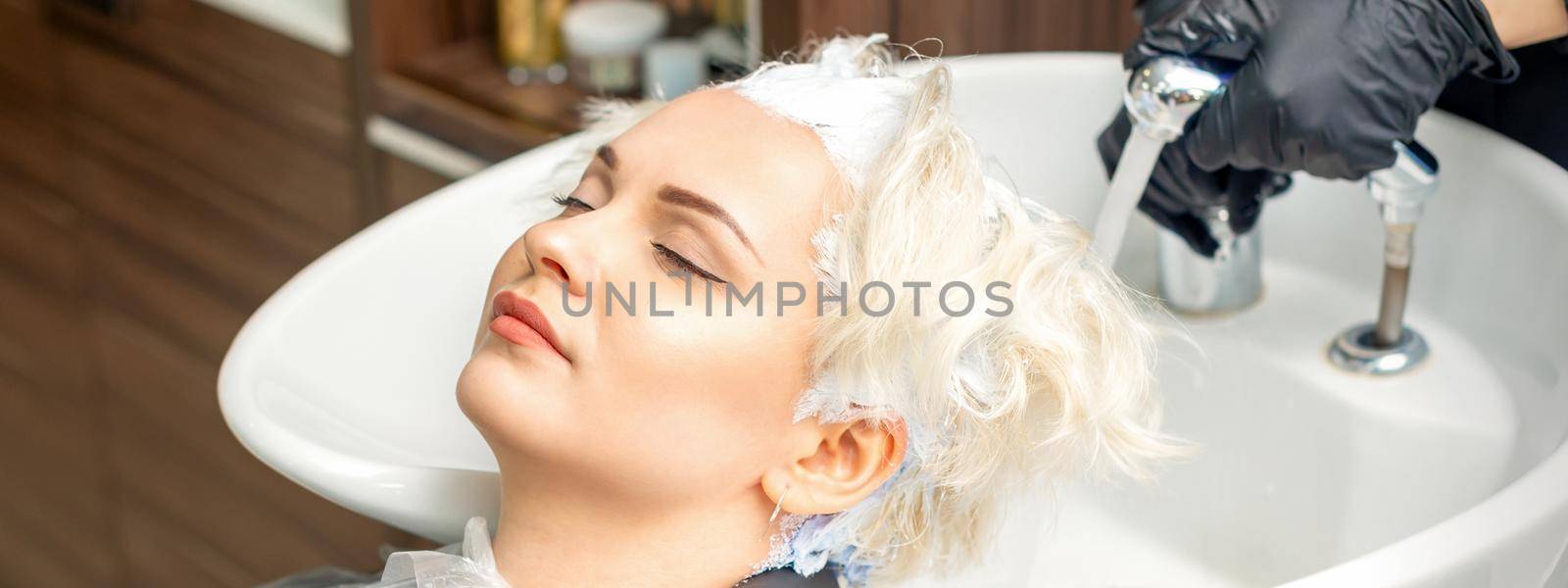 Hairdresser prepare to washes off the white dye from hair of young caucasian woman in sink at beauty salon