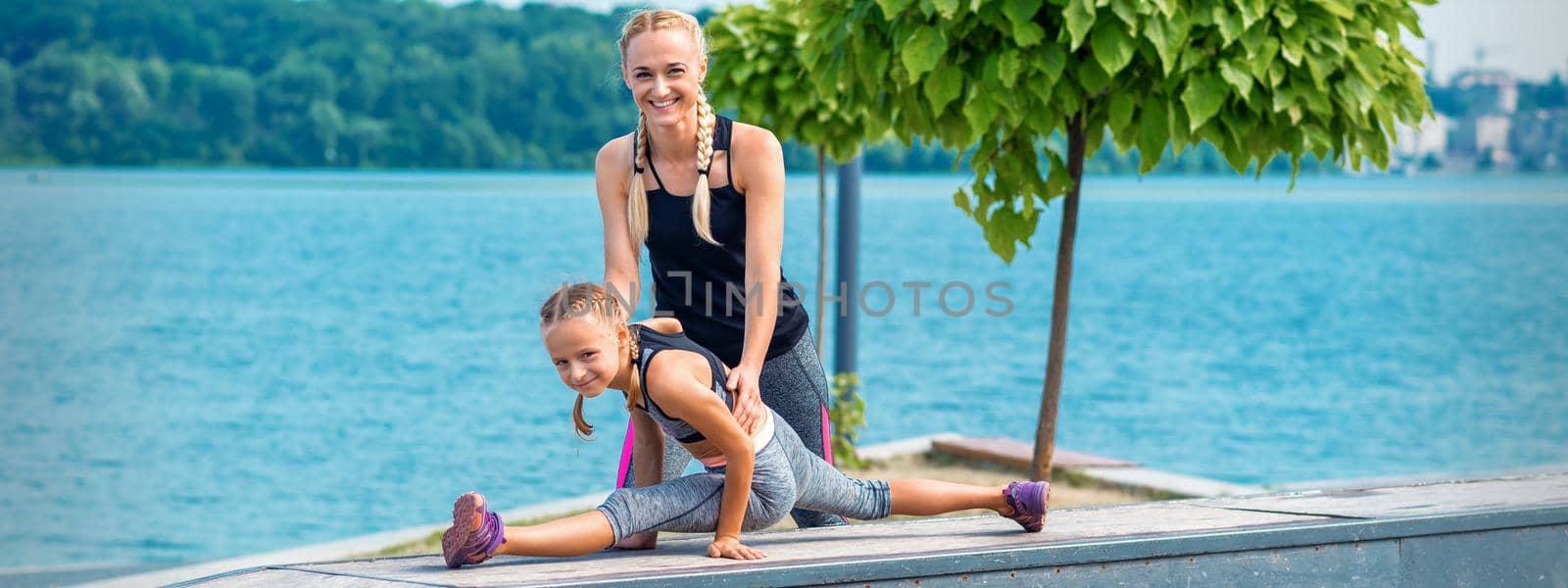 Mother and daughter doing gym exercises on the grass at the pier of the river