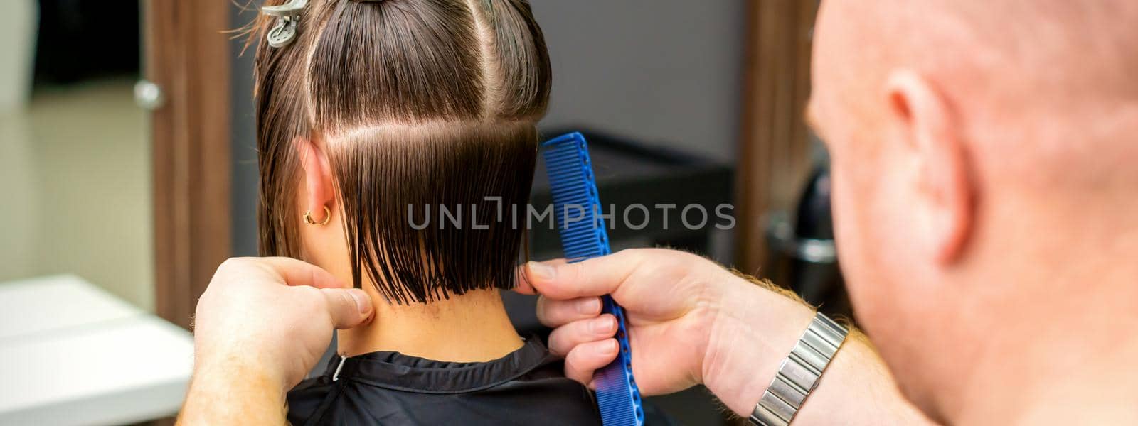 Male hairdresser cutting hair of young woman holding comb at hair salon. Rear view