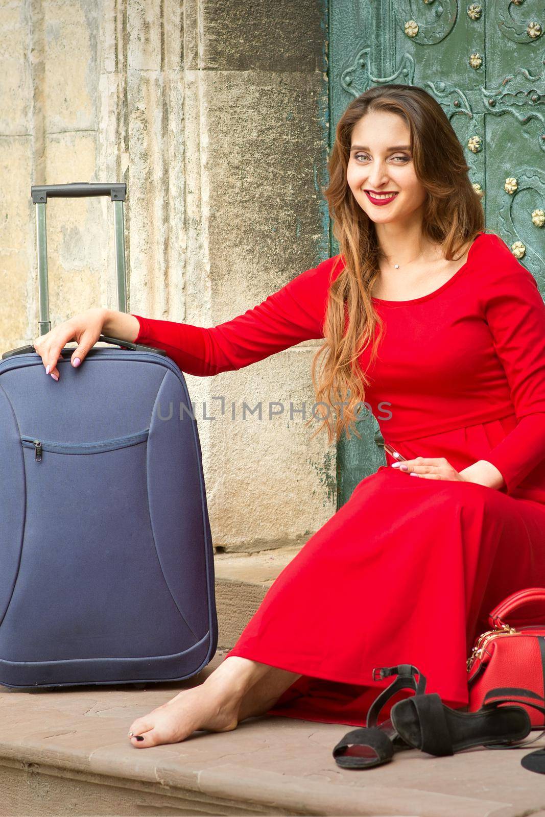 Beautiful young caucasian woman sitting on the stairs by the door wearing black hat and long red dress with travel suitcase and smartphone