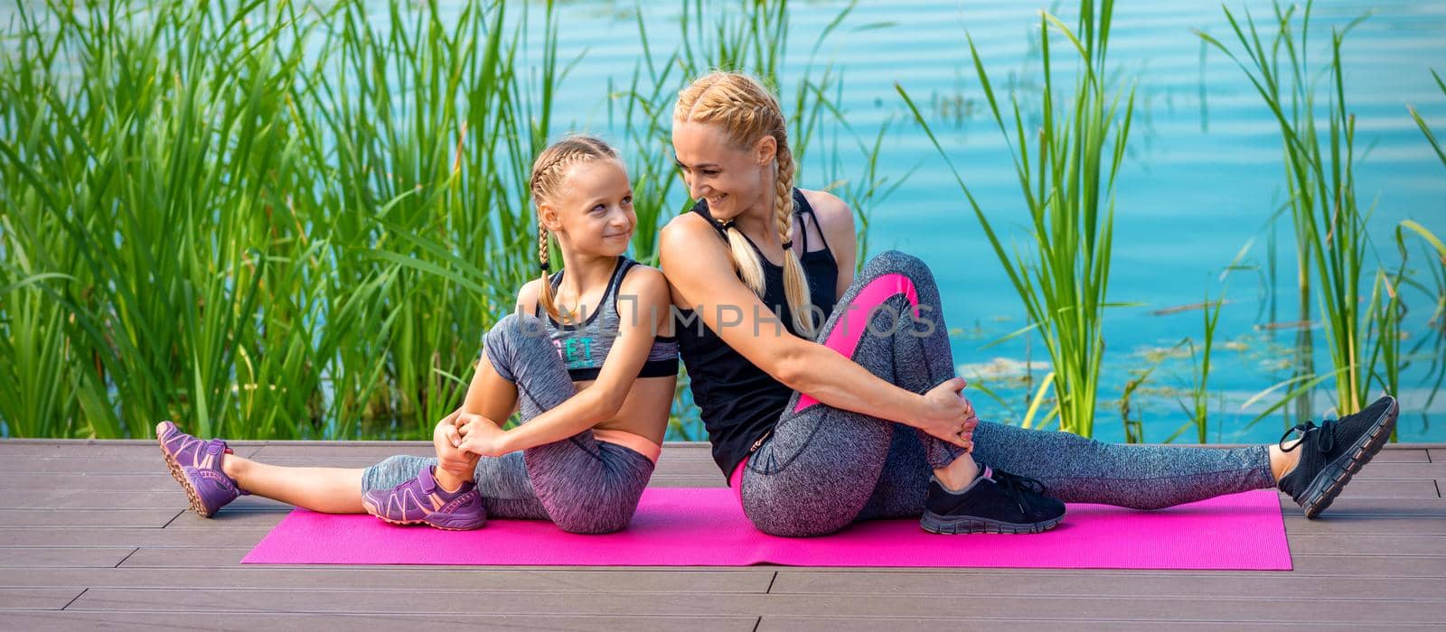 Mother and daughter doing gym exercises on the grass at the pier of the river