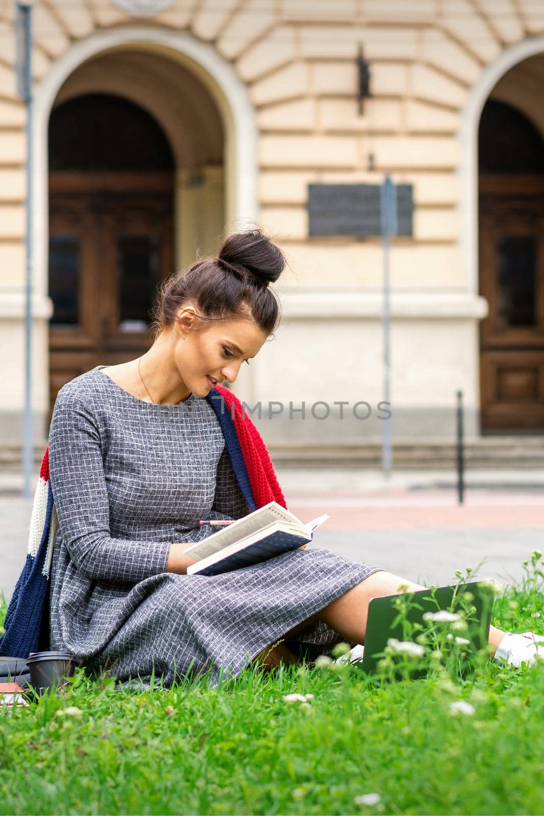 Young caucasian student woman reading the book sitting on the grass near university on the street