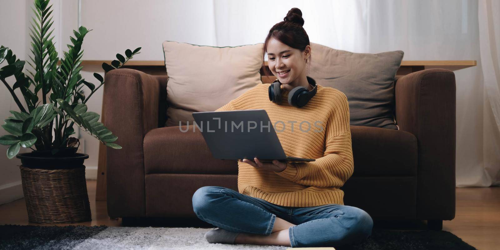 Smiling young woman working on laptop sitting on the floor in front of the sofa wearing headphones at home by wichayada