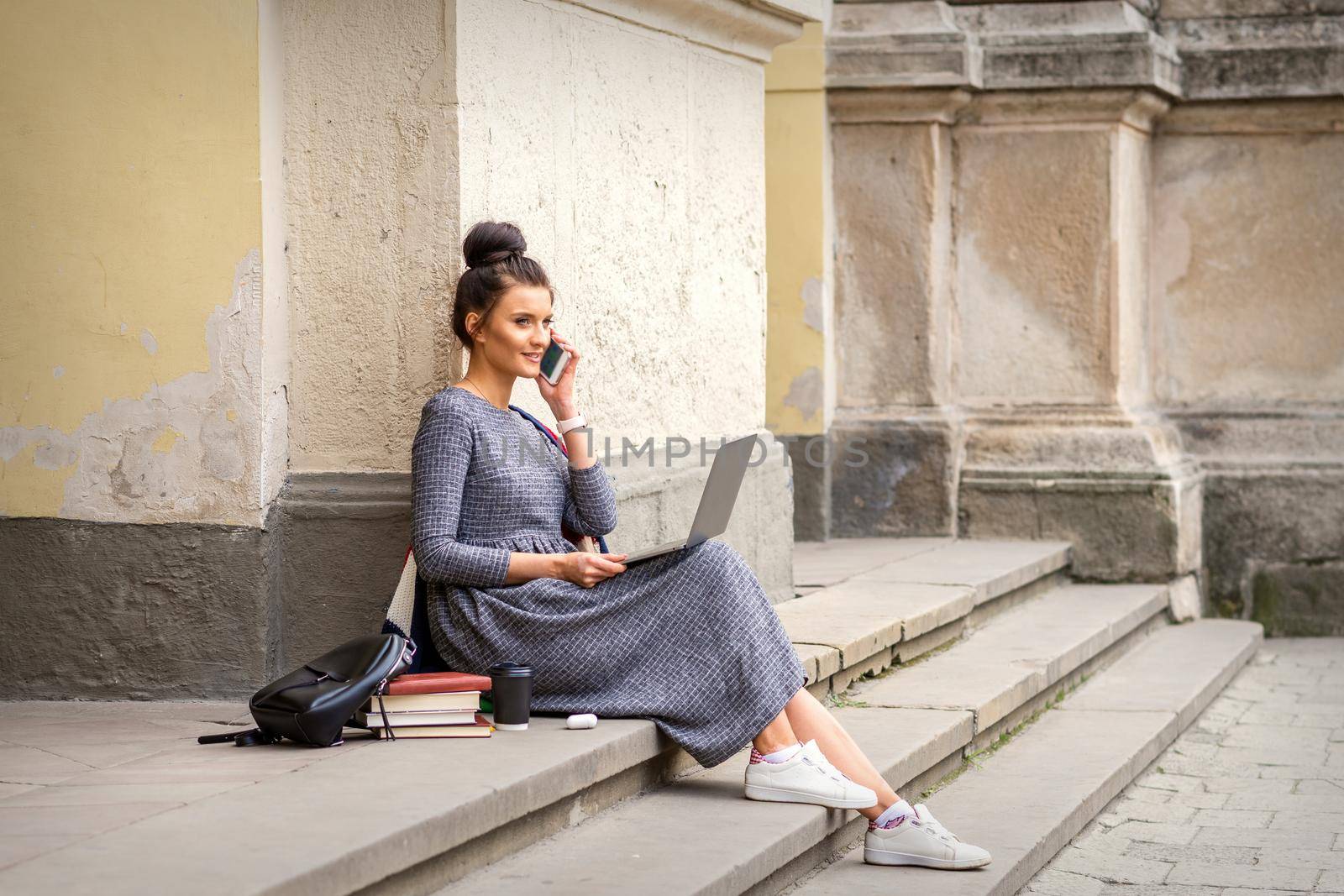 Young adult female student talking by smartphone sitting on stairs with laptop near university building