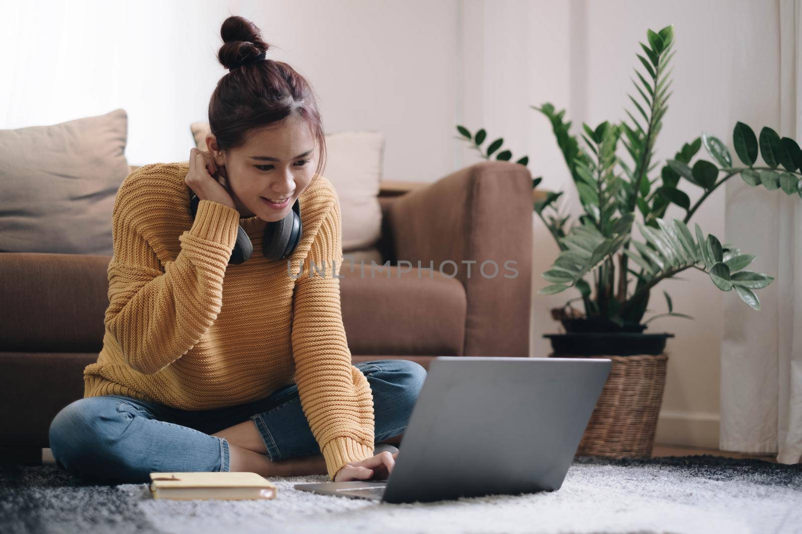 Smiling young woman working on laptop sitting on the floor in front of the sofa wearing headphones at home.