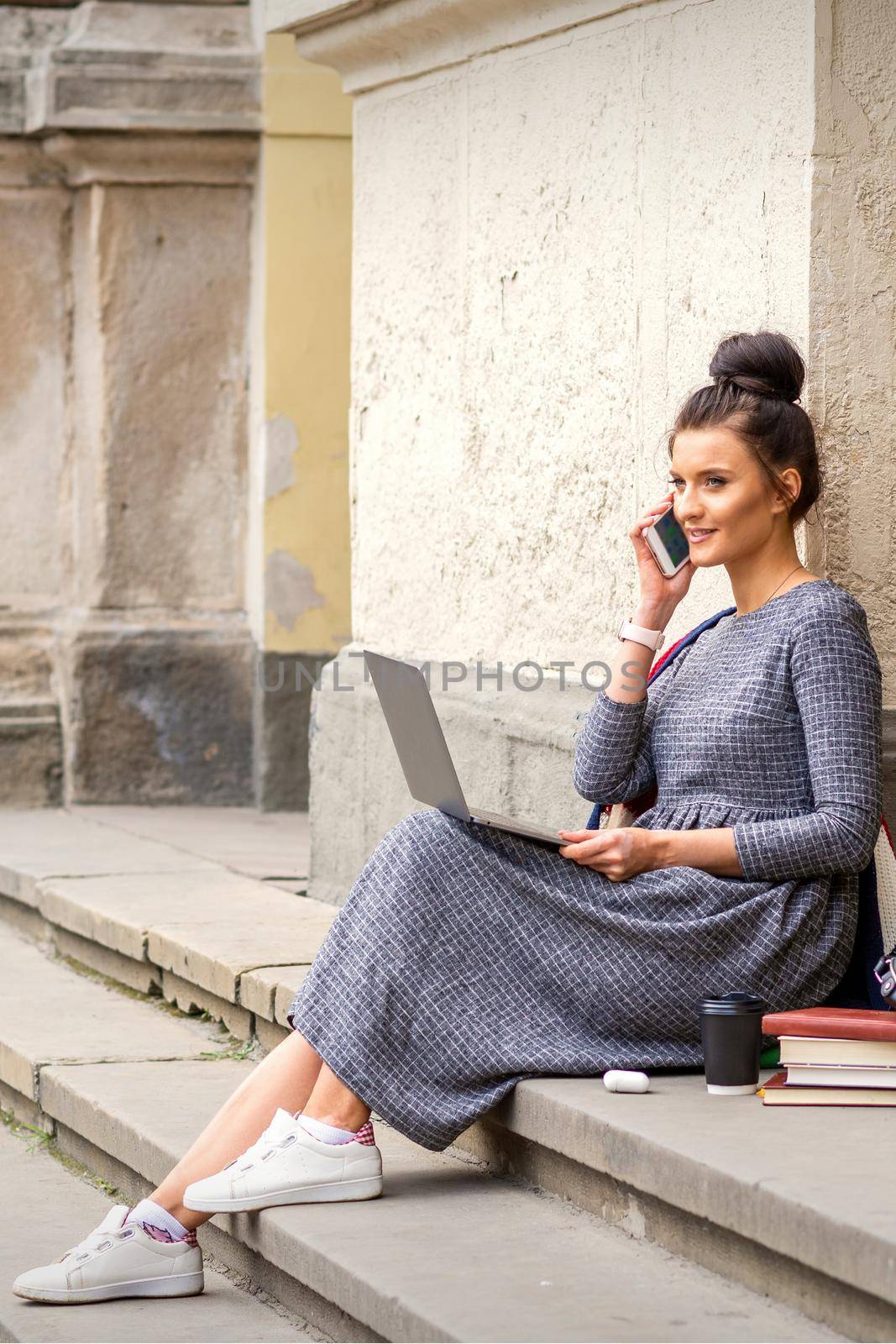 Young adult female student talking by smartphone sitting on stairs with laptop near university building
