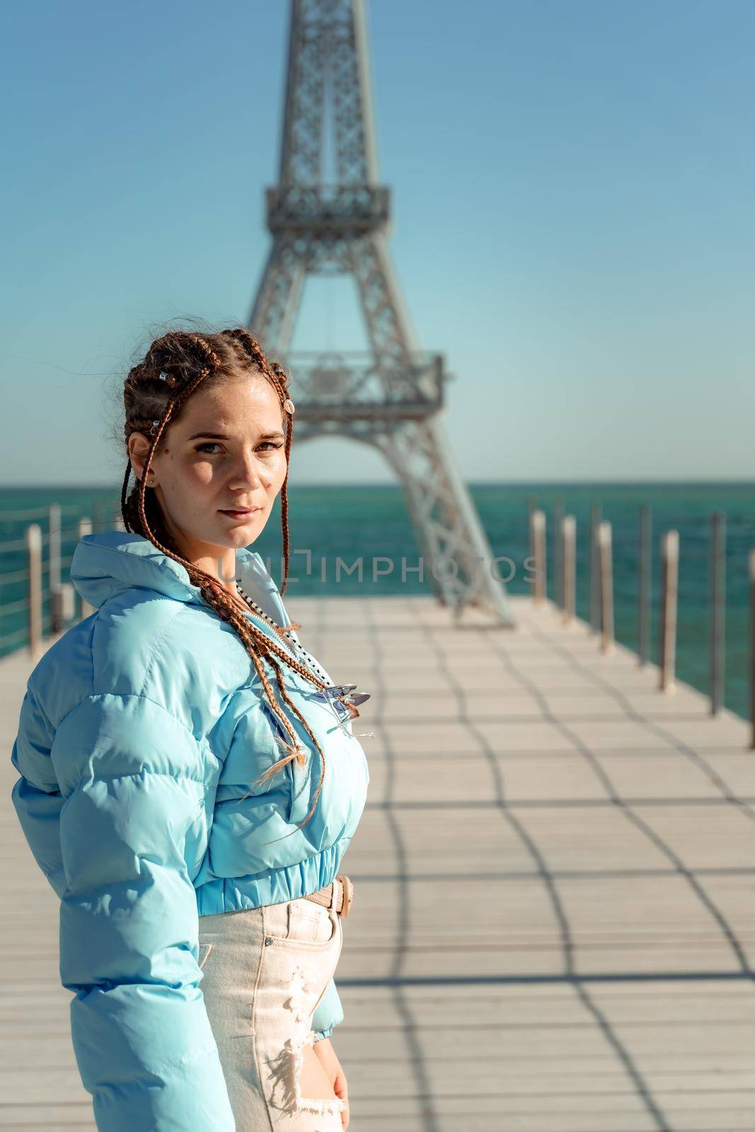 Large model of the Eiffel Tower on the beach. A woman walks along the pier towards the tower, wearing a blue jacket and white jeans